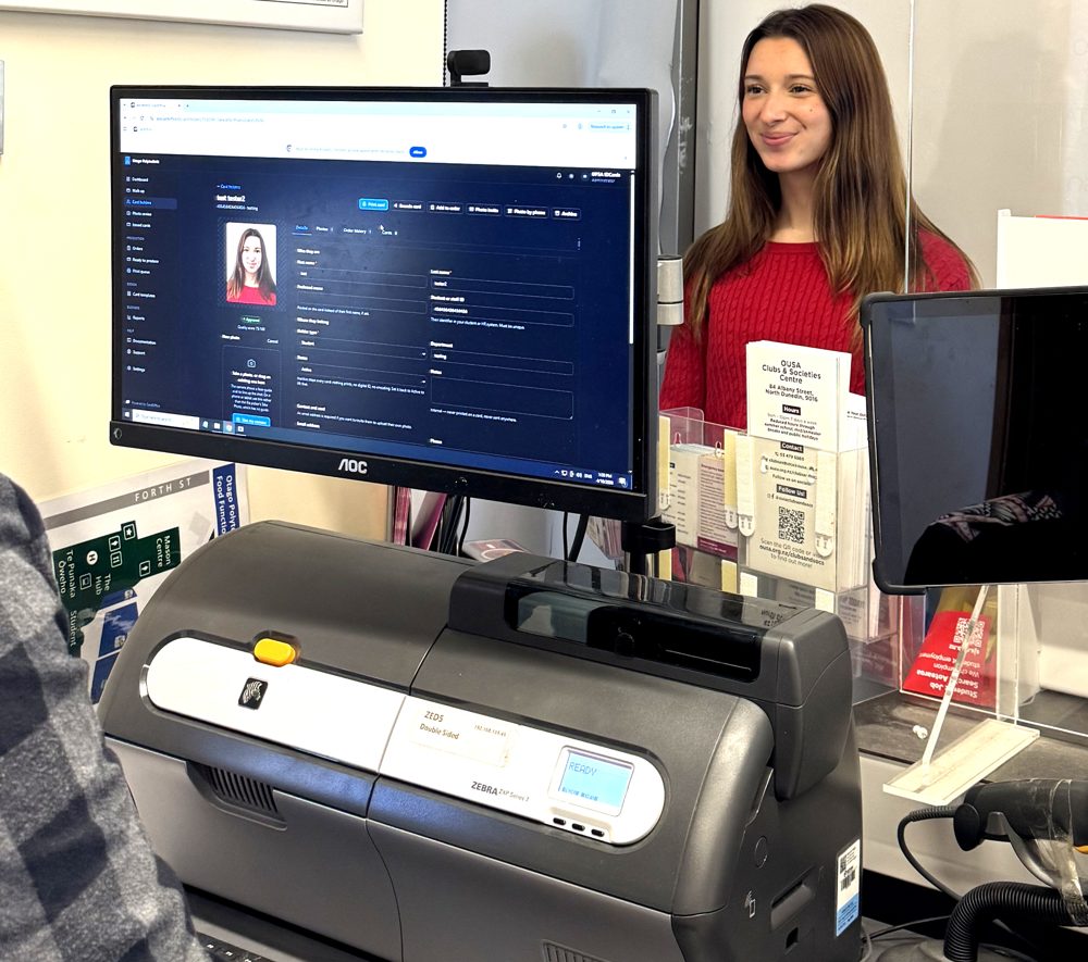 A card office counter: the CardOffice cardholder screen showing an approved photo, a card printer beside it, and a smiling cardholder waiting behind the counter